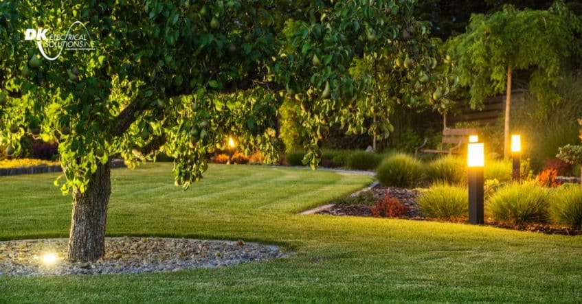 A landscaped garden at dusk features illuminated pathway lights, neatly trimmed grass, and various trees and shrubs—showcasing the beauty and value of installing outdoor lighting without exceeding your outdoor lighting cost expectations.