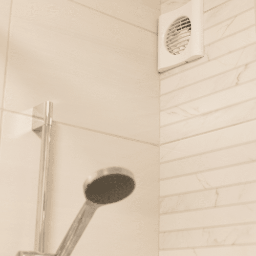 A shower head and a wall mounted bathroom fan are positioned in the corner of a tiled bathroom, ensuring proper ventilation.