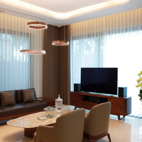 Modern living room with brown leather sofa, two beige chairs, a marble coffee table, TV on a wooden stand, circular pendant lights, and large windows with sheer curtains—explore the cost to install recessed lighting for a seamless look.