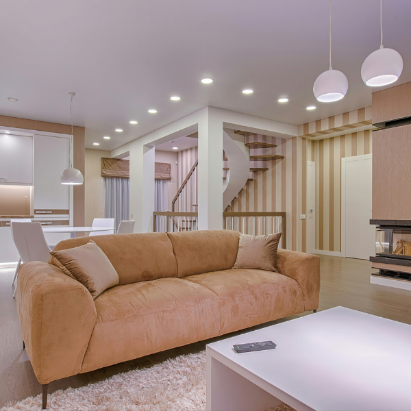 Modern living room with a tan sofa, white coffee table, and open kitchen-dining area; spiral staircase and pendant lights complement recessed lighting installation in this stylish, neutral-toned space.