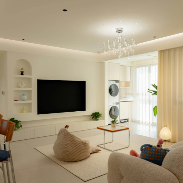 Modern living room with a large wall-mounted TV, bean bag chair, small table, indoor plants, and a stacked washer and dryer in an adjacent area. Neutral colors and soft lighting enhance the space—consider the cost to install recessed lighting for added ambiance.