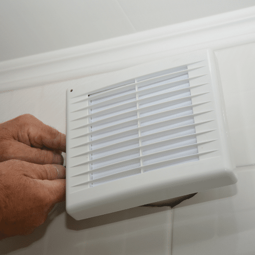 A person performs a wall mounted bathroom exhaust fan installation, adjusting a white ventilation fan or air vent cover on a wall with white tiles.