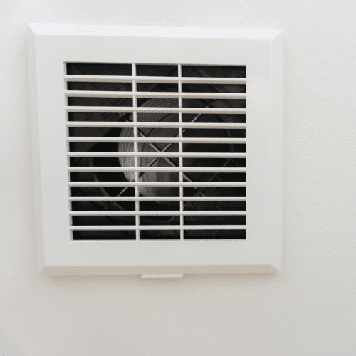 A white, square wall mounted exhaust fan with horizontal slats is expertly installed on a plain, light-colored wall.