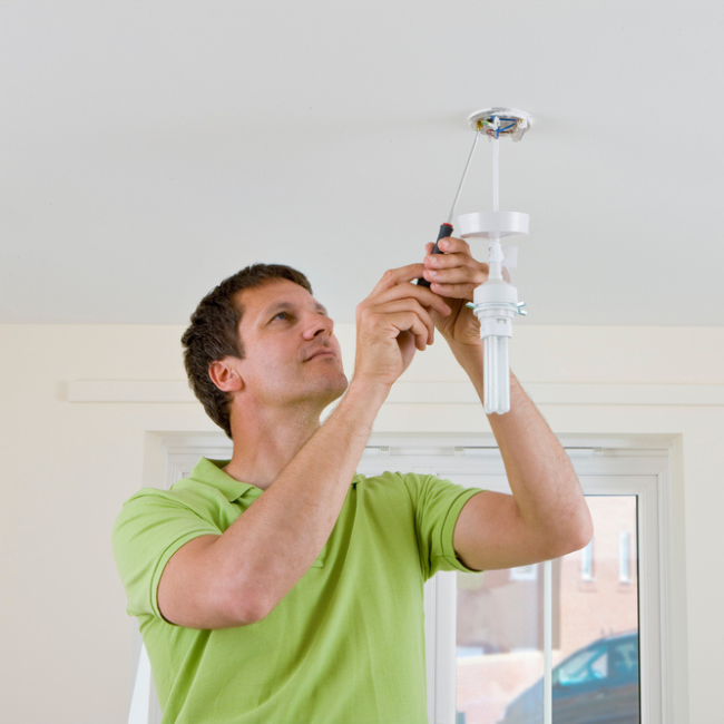 A residential electrician in a green polo shirt installs or repairs a ceiling light fixture with a screwdriver in a well-lit room, offering professional electrician services in Hamilton NJ.