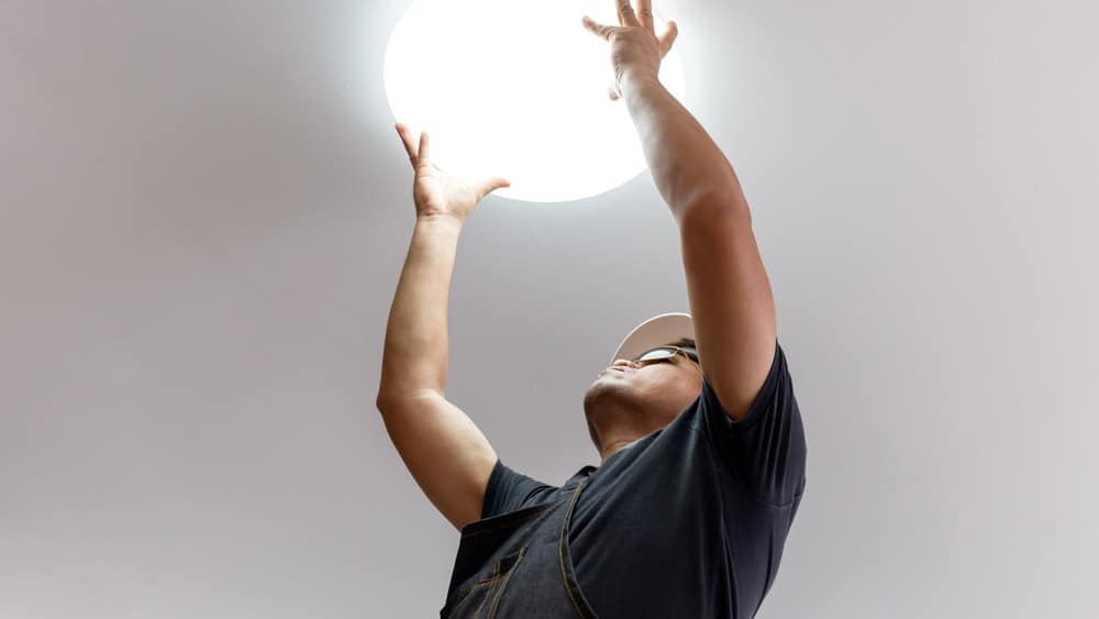 Person wearing a cap and work clothes replacing or adjusting a round ceiling light fixture, viewed from a low angle—perfect example of the reliable local electrical solutions offered by Electricians Pemberton NJ.