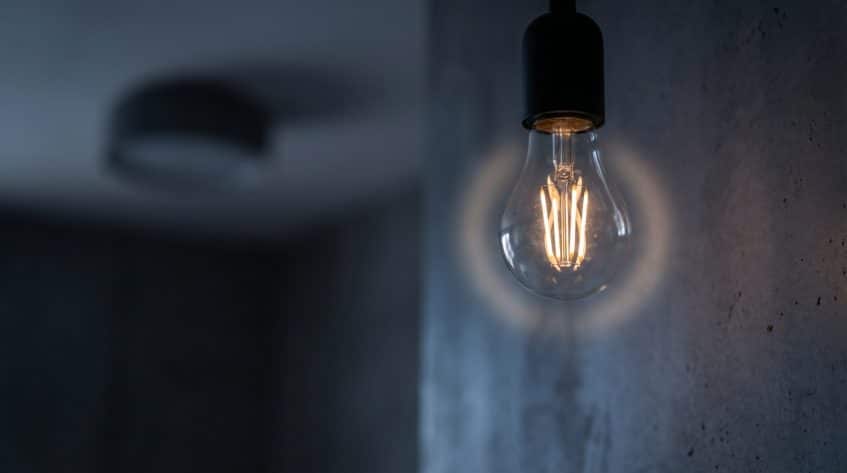 A lit incandescent light bulb hangs from the ceiling, casting a soft glow on a gray, textured wall—subtly hinting at possible house electrical issues or flickering lights in the dimly lit room.